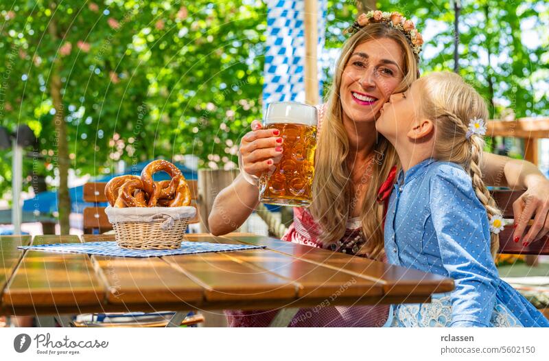 Tochter küsst ihre Mutter in Tracht, Dirndl auf die Wange und lächelt in die Kamera und stößt mit einem Krug Bier im bayerischen Biergarten oder auf dem Oktoberfest an