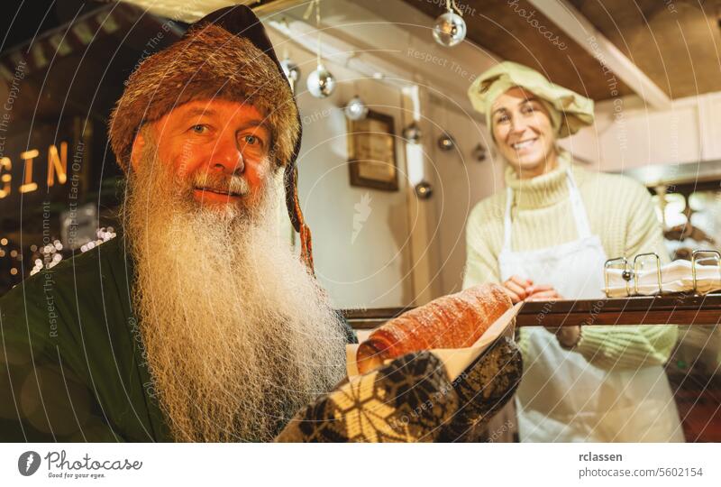 Glücklicher alter Mann mit Trdelnik, Kurtoscalacs oder Baumstriezel in der Hand in einem Stand auf dem Weihnachtsmarkt Vollbart Dezember Ungarn Brot Laden