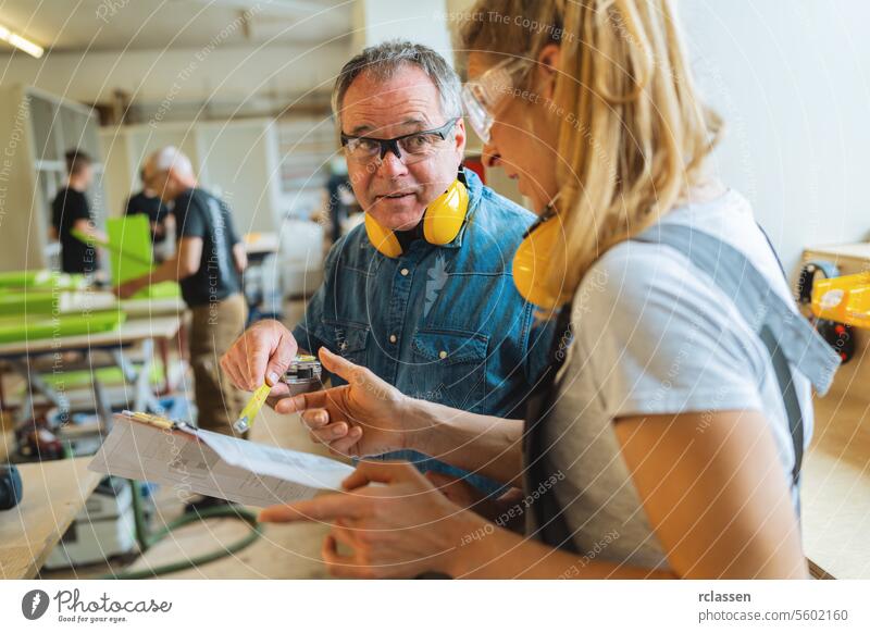 Ausbilder erklärt einem Lehrling in einer Tischlerei die Maße auf einem Bauplan professioneller Handwerker Teamwork Hobelbank Möbelindustrie Arbeiter