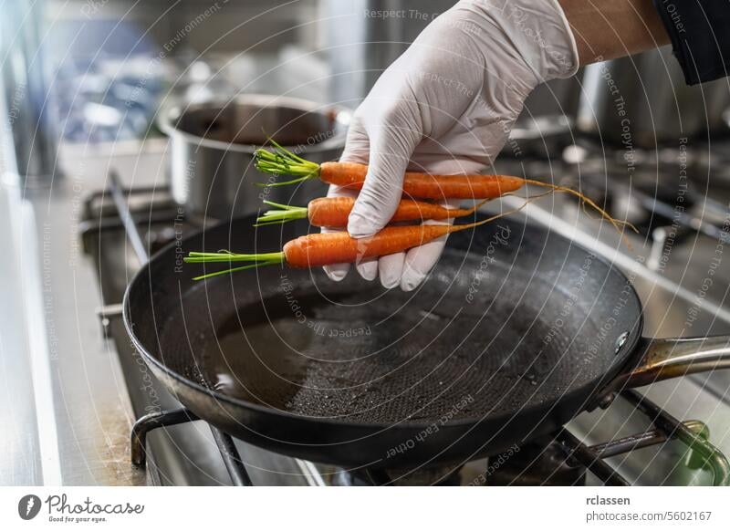 Hand legt frische Karotten in eine heiße geölte Pfanne auf einem Gasherd in einer professionellen Küche in einem Restaurant. Luxus-Hotel Kochen Konzept Bild.