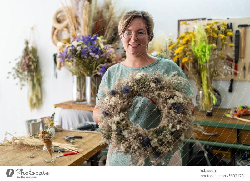 Frau mit Brille, die lächelnd einen großen Trockenblumenkranz in einem Handwerksbetrieb hält Lächeln Kranz aus Trockenblumen Floristik Dekoration & Verzierung