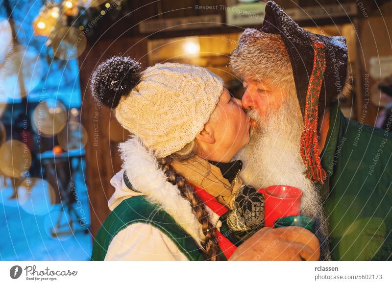 Älteres Paar, das sich auf dem Weihnachtsmarkt küsst, mit roten Tassen in der Hand, Bokeh-Lichter im Hintergrund Wintermode Tourist Frohe Weihnachten Kakao