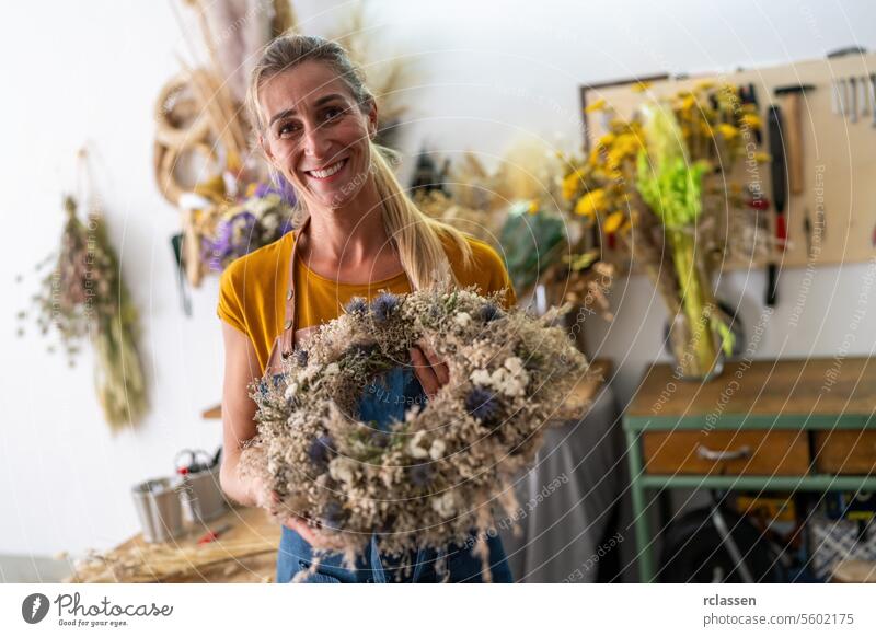 Fröhliche Floristin, die einen fertigen Trockenblumenkranz in einer Werkstattumgebung hält Blumenhändlerin Kranz aus Trockenblumen freudig Blumendesign