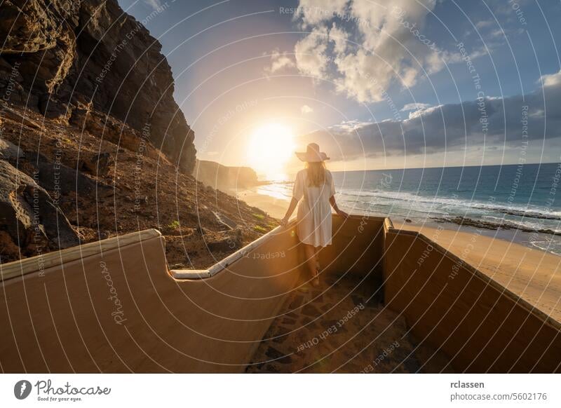 Frau mit Hut steigt bei Sonnenuntergang eine Strandtreppe hinunter, mit Blick auf den Ozean am Playa de Cofete, Fuerteventura, Kanarische Inseln. Cofete Strand