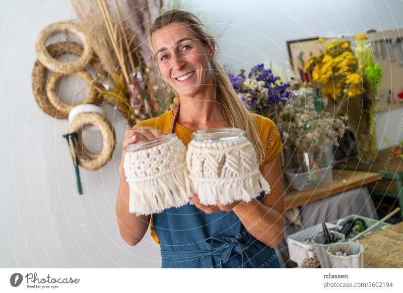 Fröhliche Floristin hält mit Makramee verzierte Gläser in ihrer Kreativwerkstatt Blumenhändler freudig Lächeln Frau Basteln handgefertigt Dekor diy