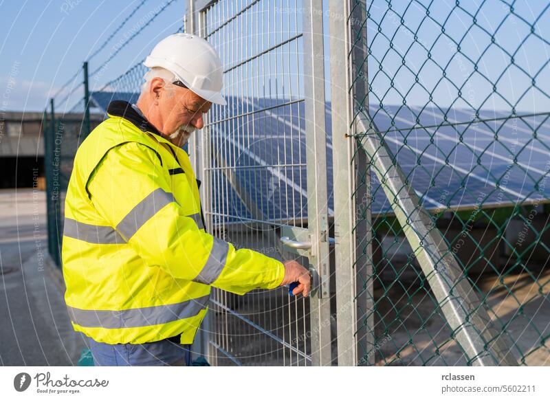 Elektroingenieur in Schutzkleidung beim Öffnen eines Tores zu einem Solarfeld Elektrotechniker Fangvorrichtung öffnendes Tor reflektierende Weste