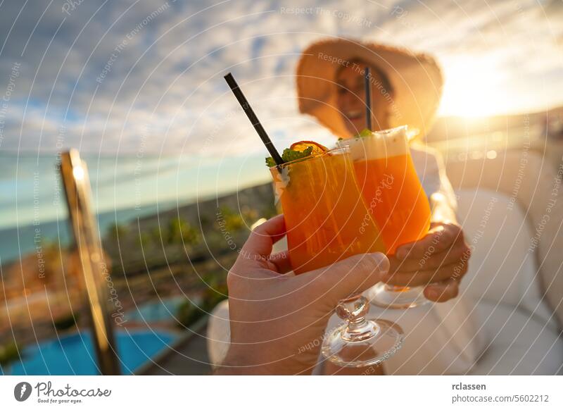 Nahaufnahme eines tropischen Cocktails mit einem Strohhalm in der Hand einer Frau mit Sonnenhut in einem tropischen Strandhotel bei Sonnenuntergang