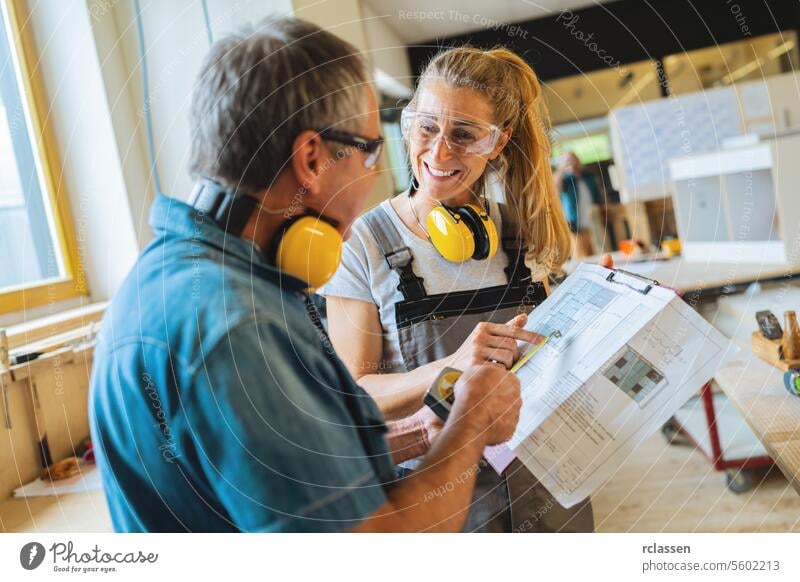 Zwei Tischler besprechen ein Projekt mit einem Bauplan in einer hellen Tischlerei professioneller Handwerker Teamwork Hobelbank Möbelindustrie Arbeiter