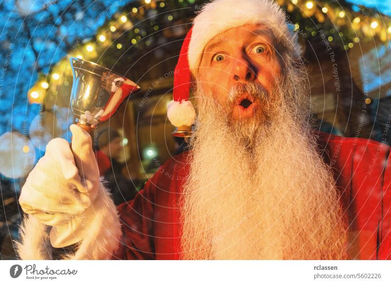 Weihnachtsmann mit überraschtem Blick, der auf einem Weihnachtsmarkt eine Glocke läutet, festlicher Lichterhintergrund Tourist freudig Frohe Weihnachten