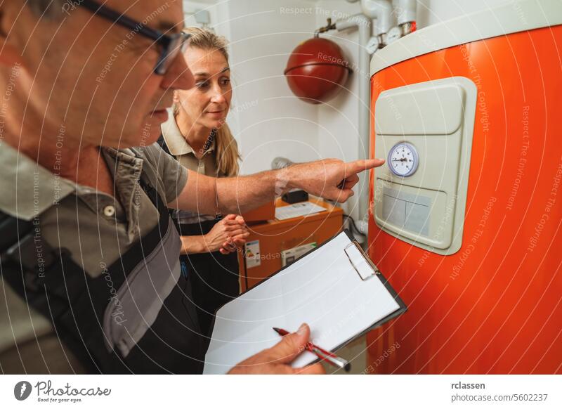 Ein Team von Heizungsmonteuren überprüft ein altes Gasheizungsthermostat und hält ein Klemmbrett mit einer Checkliste in einem Heizungsraum in einem Haus. Gasheizung Ersatz Verpflichtung Konzept Bild