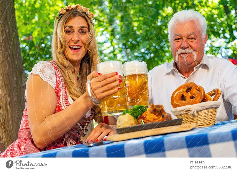 Bayerische Frauen und ein alter Mann stoßen mit Bierkrügen an und genießen traditionelle bayerische Küche mit gebratenen Schweinshaxen in einem Biergarten oder auf dem Oktoberfest