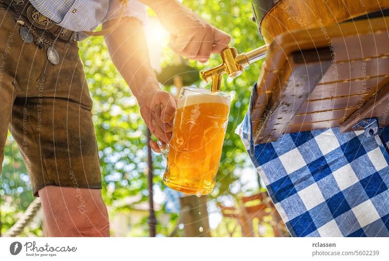 Ein bayerischer Mann in Lederhosen schenkt ein großes Lagerbier aus einem hölzernen Bierfass im Biergarten ein. Hintergrund für Oktoberfest oder Wiesn, Volks- oder Bierfest