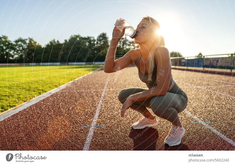 Frau trinkt Wasser aus einer Wasserflasche, während sie bei Sonnenuntergang auf einem Sportplatz hockt Trinkwasser Hydratation Leichtathletik Fitness Athlet