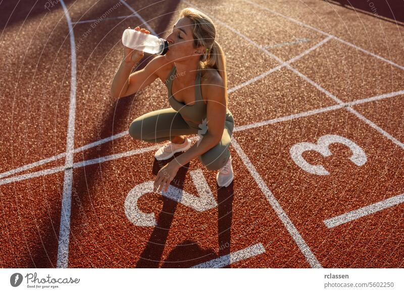 Sportlerin, die Wasser trinkt, während sie bei Sonnenuntergang auf einem Sportplatz hockt Athlet Frau Trinkwasser hockend Leichtathletik Hydratation Fitness