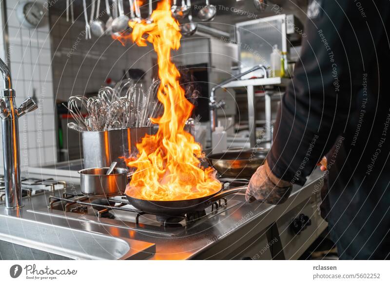 Profikoch flambiert in der Küche in einer Pfanne auf einem gasbefeuerten Herd. Luxus-Hotelkoch Kochen Konzept Bild. Flamme Küchenchef Essen zubereiten