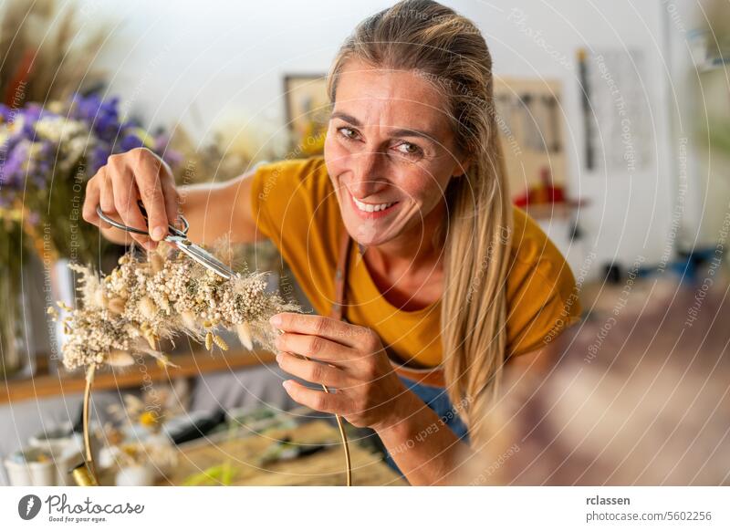 eine lächelnde Floristin schneidet ein zartes Trockenblumenarrangement zurecht, während sie in einem mit floralen Elementen gefüllten Atelier akribisch arbeitet