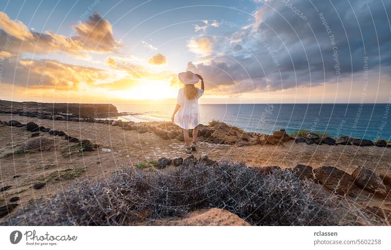 Rückansicht einer Frau in Weiß mit Hut und Blick auf das Meer während des Sonnenuntergangs an der felsigen Küste von Playa de Cofete, Fuerteventura, Kanarische Inseln.