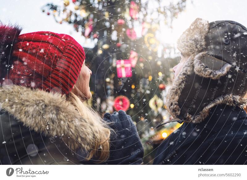 Frau mit roter Mütze und Mann mit Pelzmütze blicken auf einen Weihnachtsbaum auf dem Weihnachtsmarkt, Schneeflocken fallen an einem Wintertag, mit Platz für Ihren individuellen Text.