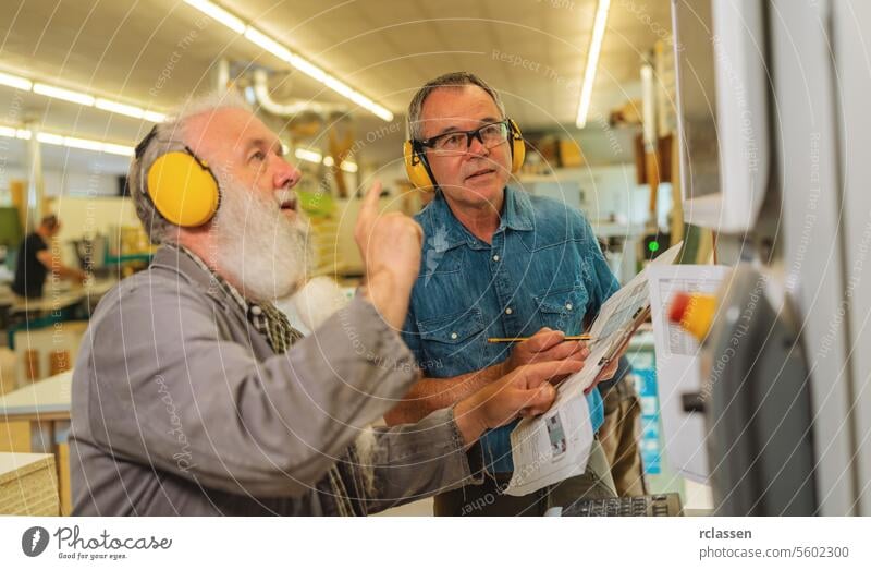 Zwei Männer mit Gehörschutz besprechen die Arbeit an einer Schneidemaschine in einer geschäftigen Schreinerei Maschine Tischlerwerkstatt Diskussion über