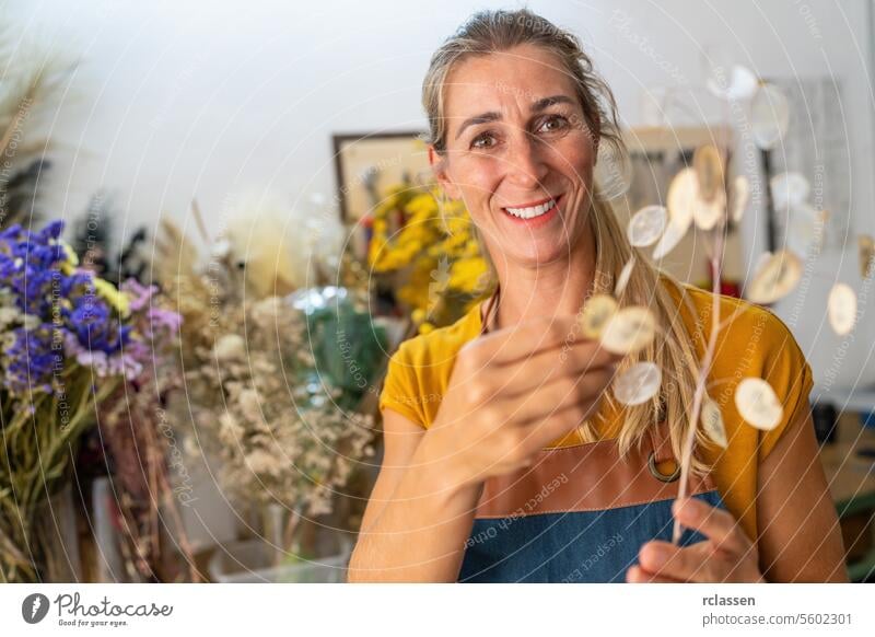 Fröhliche Blumenhändlerin, die einen Zweig getrockneter Lunarien in einer Werkstatt mit unscharfen Blumen hält freudig getrocknete Lunaria verschwommene Blumen