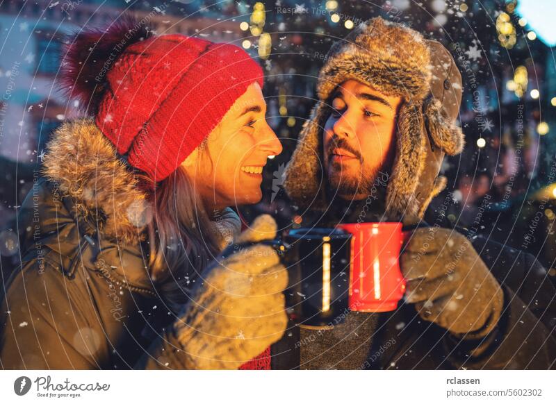 Ein Paar stößt mit Glühwein auf einem Weihnachtsmarkt an Bowle Tasse Kakao Handschuhe traditionell Deutsch Schneeflocken Feiertag Weihnachten Lichter Bokeh Wein