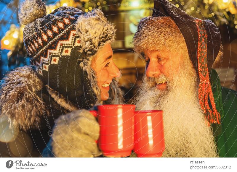 Freunde stoßen mit Glühwein- oder Kakaobechern auf dem Weihnachtsmarkt an, Frau mit Wintermütze, Mann mit weißem Bart Bowle Senior Frohe Weihnachten Tasse