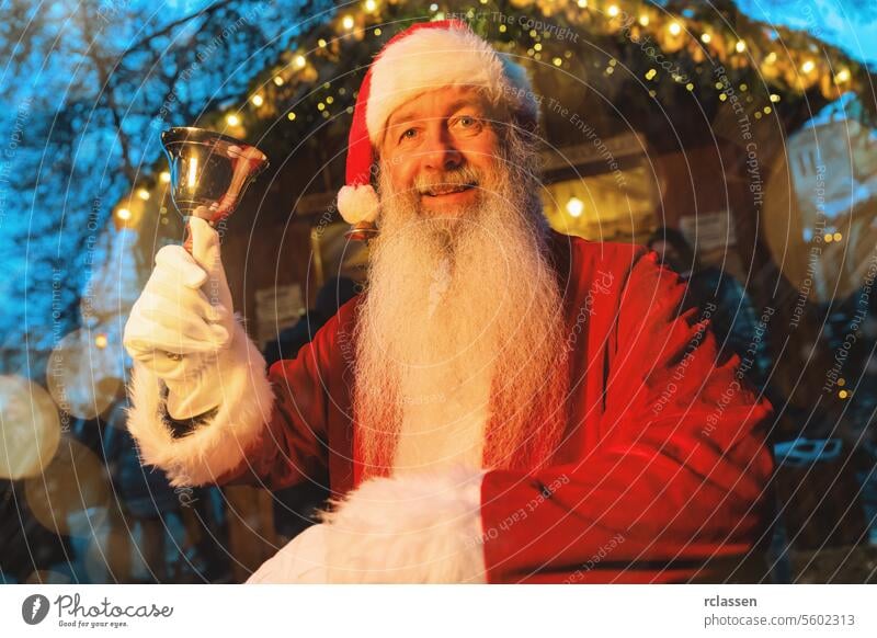 Der Weihnachtsmann hält eine Glocke mit einem freudigen Gesichtsausdruck auf einem Weihnachtsmarkt mit festlicher Beleuchtung Halteglocke freudiger Ausdruck
