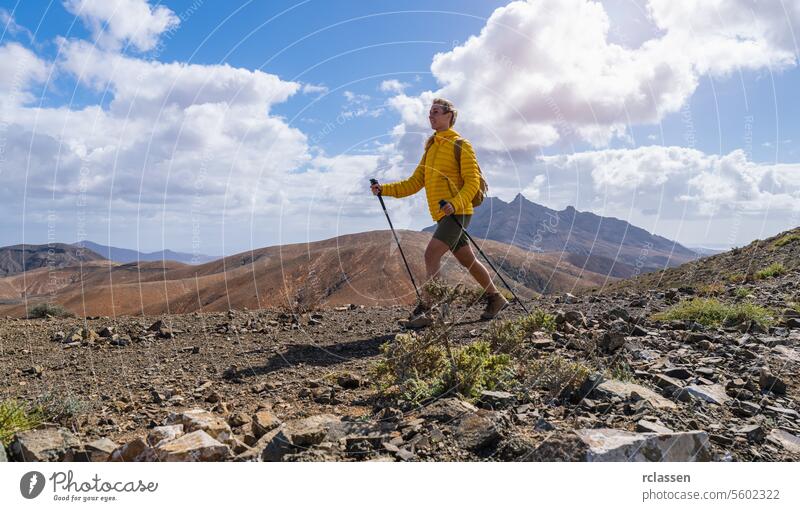 Wanderer in gelber Jacke auf Bergpfad mit bewölktem Himmel und Gipfel in der Ferne Fuerteventura gelbe Jacke Bergweg bewölkter Himmel wandern Outdoor-Erlebnis