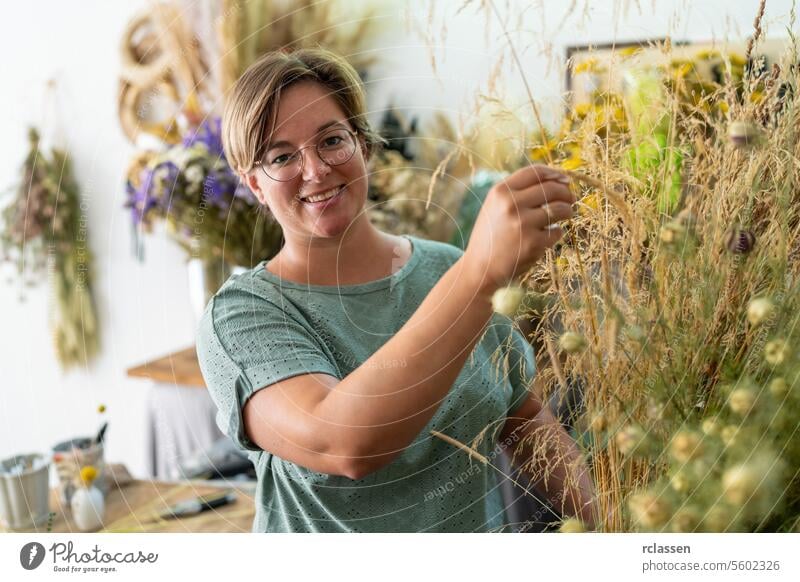 Lächelnde Frau mit Brille arrangiert getrocknete Pflanzen in einer Blumenwerkstatt mit hängenden Blumensträußen lächelnde Frau arrangierend Hängesträuße