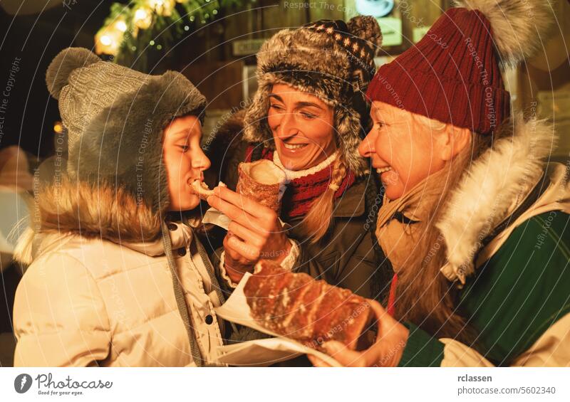 Glückliche Oma und Mutter füttern ihre Tochter mit einem Baumstriezel oder Trdelnik vor einem Weihnachtsmarkt in Deutschland Familie süß Snack Zucker Spirale