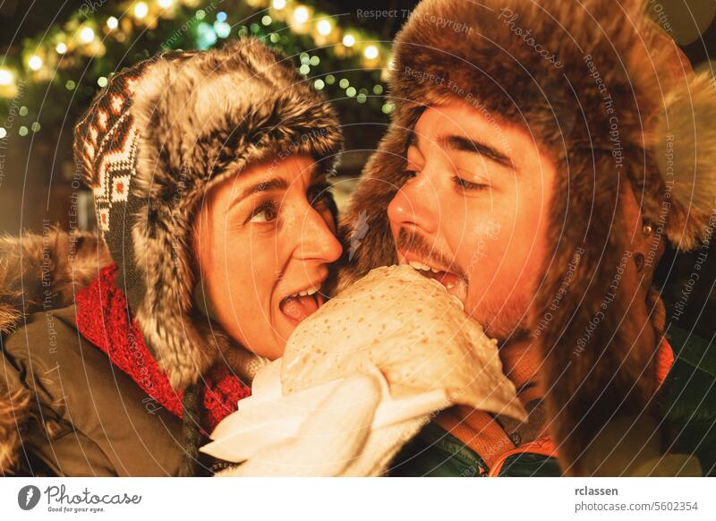 Pärchen isst spielerisch einen Crêpe auf einem Weihnachtsmarkt, beide tragen Wintermützen Schokolade Frohe Weihnachten Handschuhe traditionell Glühwein Deutsch