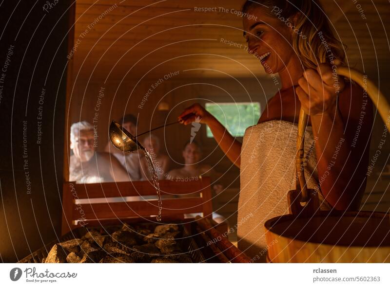 Glückliche Frau gießt Wasser auf heiße Steine im Saunaraum mit einer Gruppe von Menschen. Dampf und Wasser auf den Steinen, Spa und Wellness-Konzept, entspannen Sie in heißen finnischen Sauna.