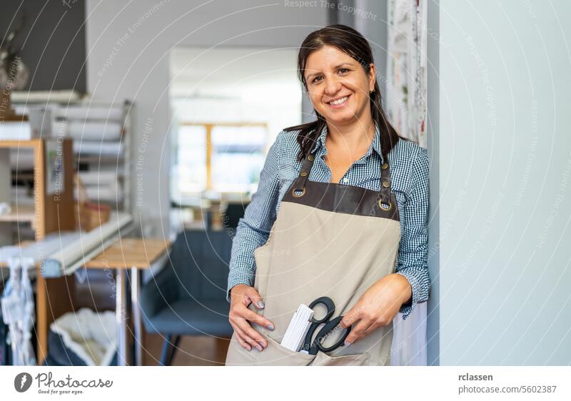 eine lächelnde Schneiderin in einem karierten Hemd und einer beigen Schürze steht in einem Arbeitsbereich und hält eine Schere und ein Maßstabslineal in ihrer Schürzentasche