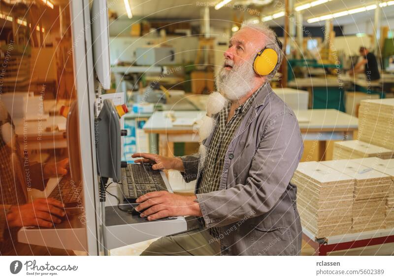 Mann mit Computer an einer Schneidemaschine in einer Holzwerkstatt Kapselgehörschützer professioneller Handwerker Hobelbank Möbelindustrie benutzend Gehörschutz