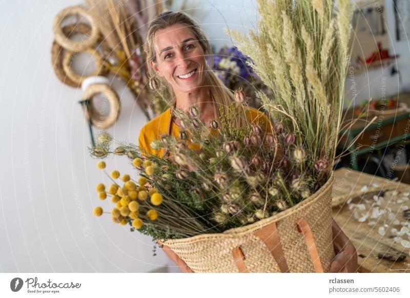 Strahlende Floristin, die einen großen Korb mit Trockenblumen in ihrem Atelier hält Blumenhändlerin strahlend großer Korb Blumenatelier Lächeln Glück Floristik