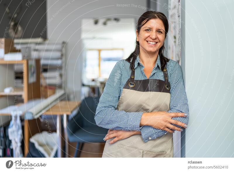 Eine selbstbewusste Frau mit kariertem Hemd und beiger Schürze steht mit verschränkten Armen in einer Werkstatt und lächelt in die Kamera. Schere Vorhänge Beruf