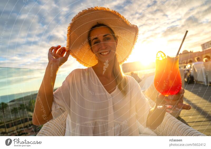 Fröhliche Frau mit Strohhut bietet einen Cocktail in einem tropischen Strandhotel mit Sonnenuntergang im Hintergrund an Paradiesstrand Playa Küstenlinie