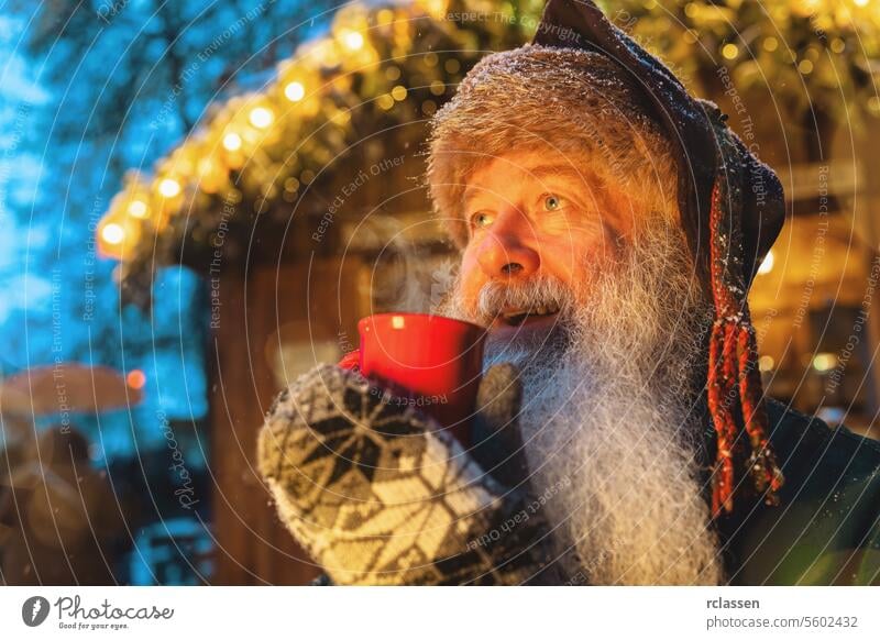 Lächelnder Mann mit Bart, der einen roten Becher mit Glühwein oder heißer Schokolade auf einem Weihnachtsmarkt hält, festliche Lichter im Hintergrund Bowle