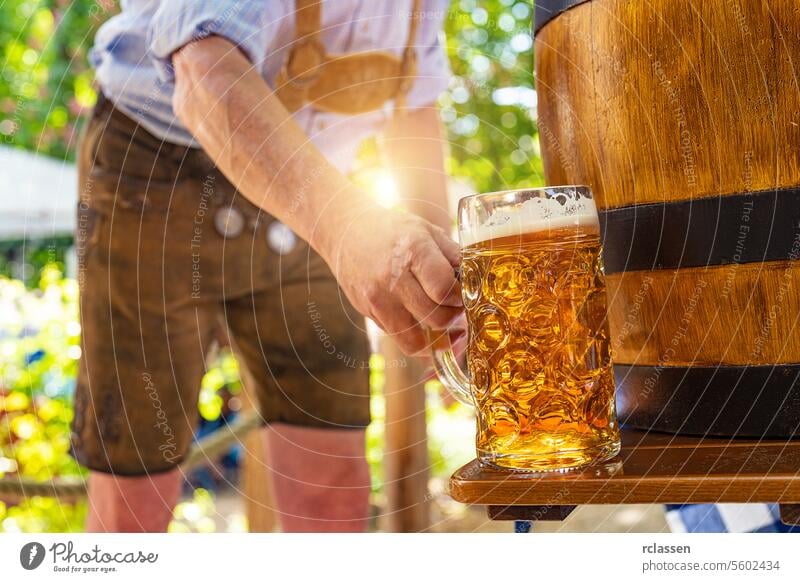 Bayerischer Mann in traditioneller Lederhose stellt frisch eingeschenktes Lagerbier auf den Tisch vor einem hölzernen Bierfass im Biergarten oder auf dem Oktoberfest