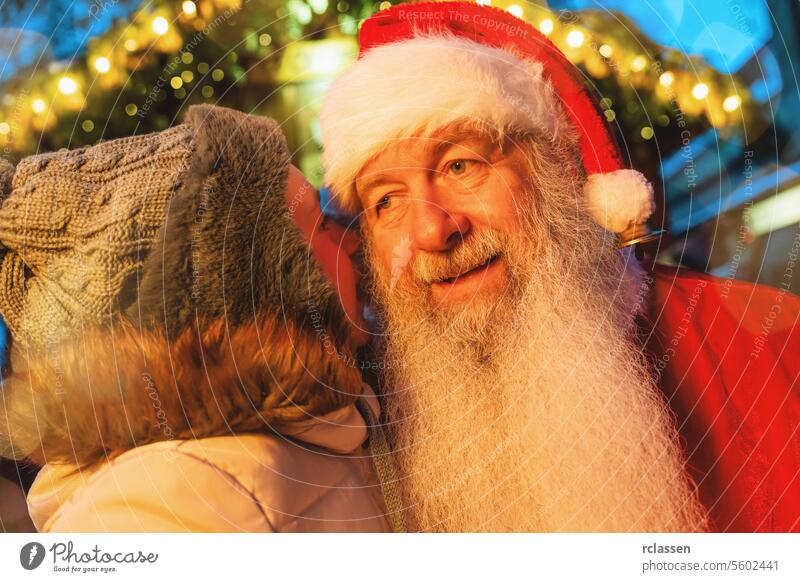 Kind flüstert dem Weihnachtsmann auf einem Weihnachtsmarkt mit festlichen Lichtern im Hintergrund zu Bokeh freudig Frohe Weihnachten Handschuhe traditionell