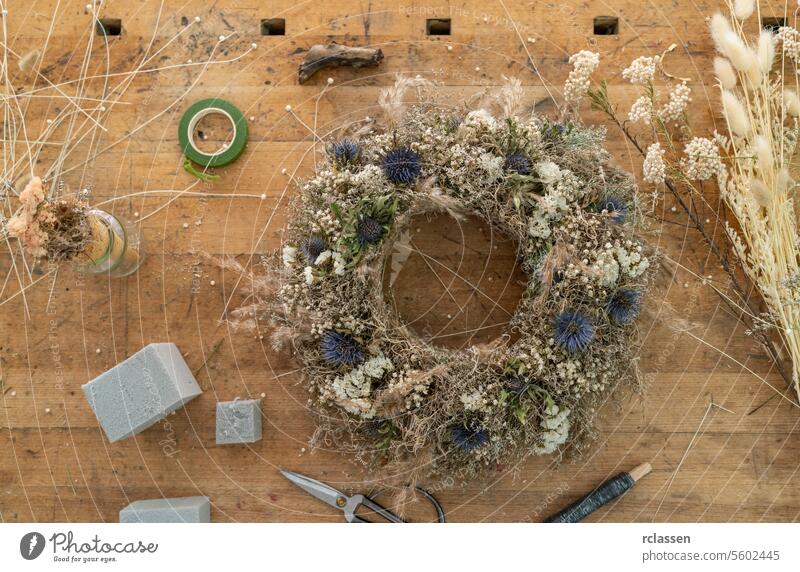 Draufsicht auf einen Blumenkranz auf einem Holztisch zwischen Floristenwerkzeugen und Bastelmaterial Ansicht von oben Trockenblumen Werkstatt diy handgefertigt