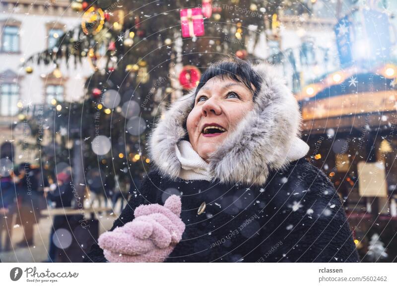 Ältere Frau im Pelzmantel mit Kapuze blickt freudig auf, als auf dem Weihnachtsmarkt Schnee fällt Tourist Frohe Weihnachten Kakao Handschuhe traditionell