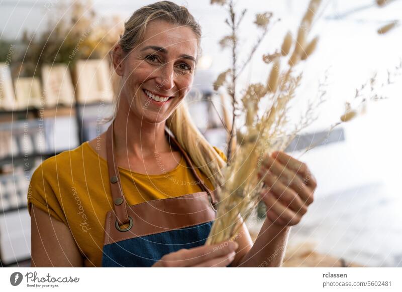 Eine lächelnde Floristin hält einen Strauß getrockneten Weizens in einer hellen Werkstatt Blumenhändlerin Lächeln Trockenweizen heiter Floristik blondes Haar