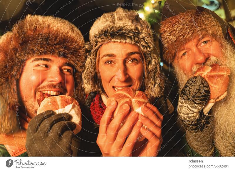 Eine Gruppe von Freunden genießt Würstchen im Brötchen auf einem lebhaften Weihnachtsmarkt, mit Wintermützen und einem Lächeln. Frohe Weihnachten Handschuhe