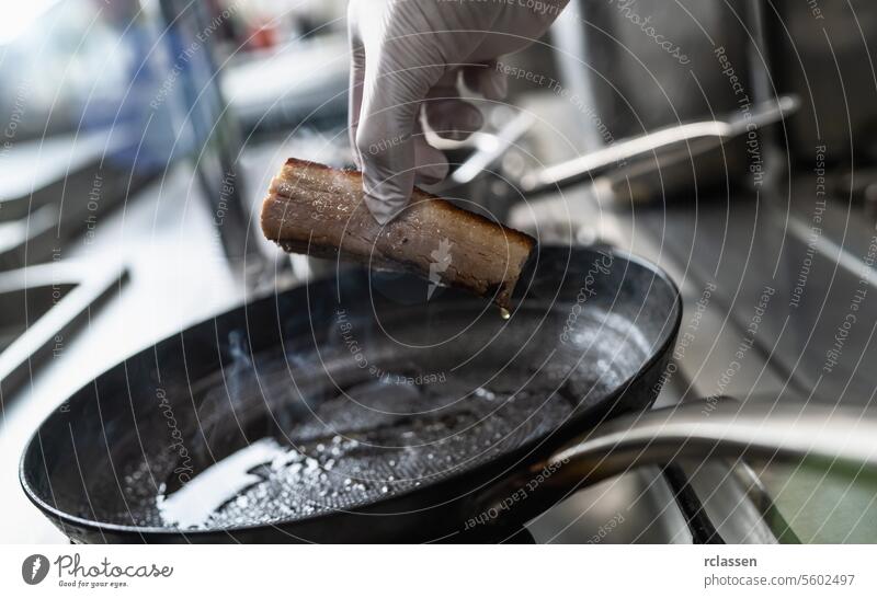 Hand hält einen knusprigen Schweinebauch, der in einer heißen geölten Pfanne auf einem Gasherd in einer professionellen Küche in einem Restaurant gebraten wurde. Luxus-Hotel Kochen Konzept Bild.
