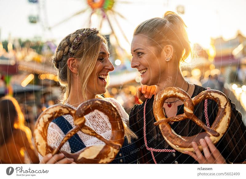 Freundinnen schauen sich an, wenn sie auf einem bayerischen Volksfest oder Oktoberfest eine Brezel oder Brezen in der Hand halten oder in Tracht oder Dirndl in Deutschland dulden