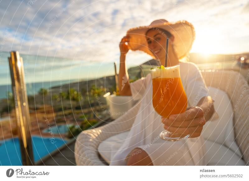 Frau mit Sonnenhut genießt einen Cocktail in einem tropischen Strandhotel auf einer sonnigen Terrasse mit einem Pool im Hintergrund Paradiesstrand Playa