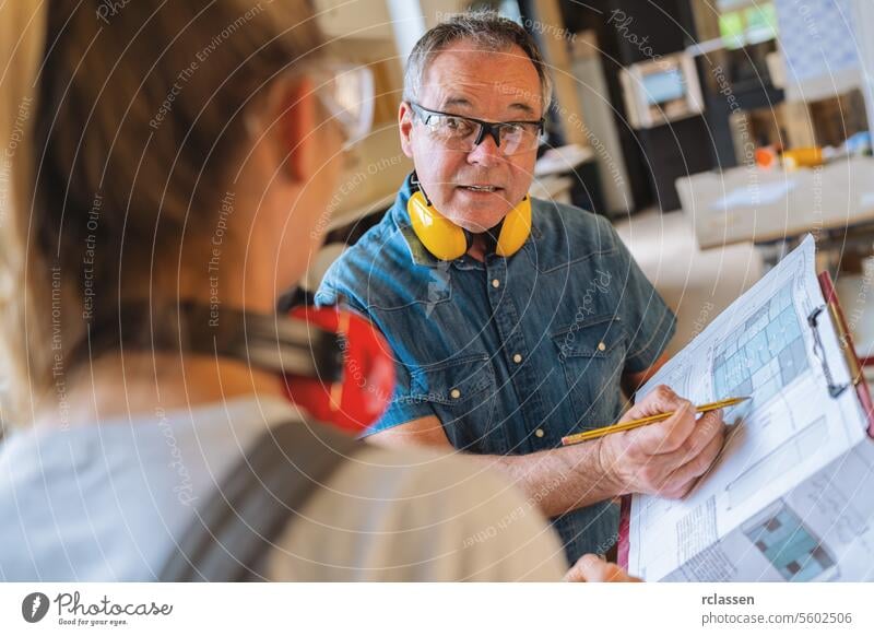 Ausbilder im Tischlerhandwerk bespricht mit einem Lehrling in einer Tischlerwerkstatt die Details eines Bauplans Teamwork professioneller Handwerker Hobelbank