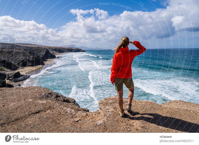 Wanderer in roter Jacke, der an einem sonnigen Tag von einer Klippe aus auf die Wellen des Meeres blickt Fuerteventura rote Jacke Meereswellen sonniger Tag