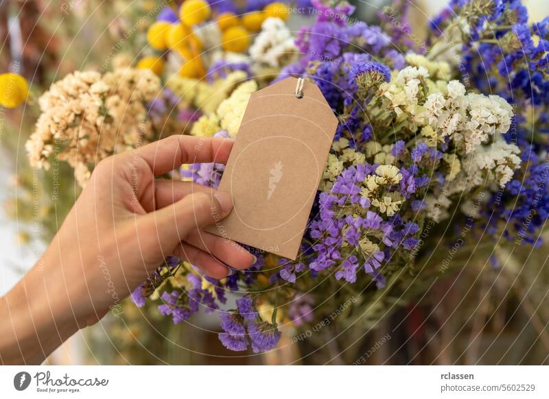 Eine Hand hält ein leeres braunes Schild vor einem Strauß bunter Trockenblumen, mit Platz für Ihren individuellen Text. dekorativ Dekor Herbst Blumenhändler